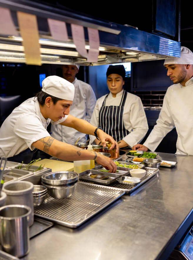 A young male chef with tattooed arms prepares a dish in a restaurant kitchen while the staff looks on