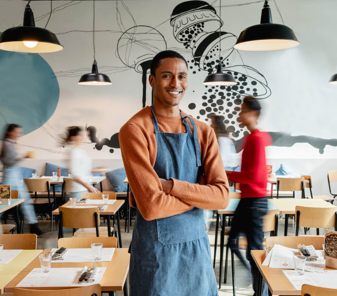 A restaurant manager standing in his restaurant smiling with folded arms with his staff walking in the background
