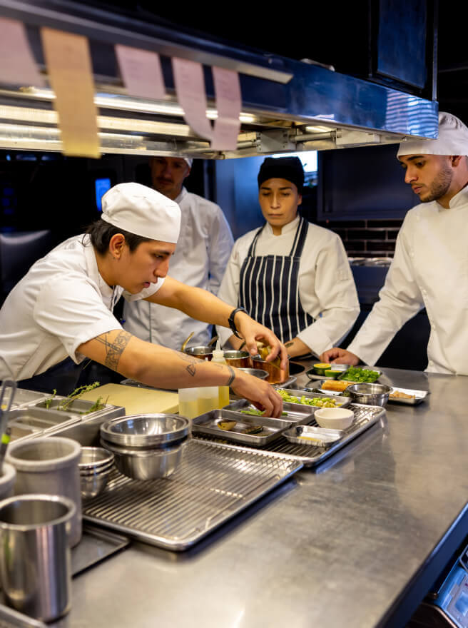 A young chef prepares a dish in a restaurant kitchen while the staff looks on