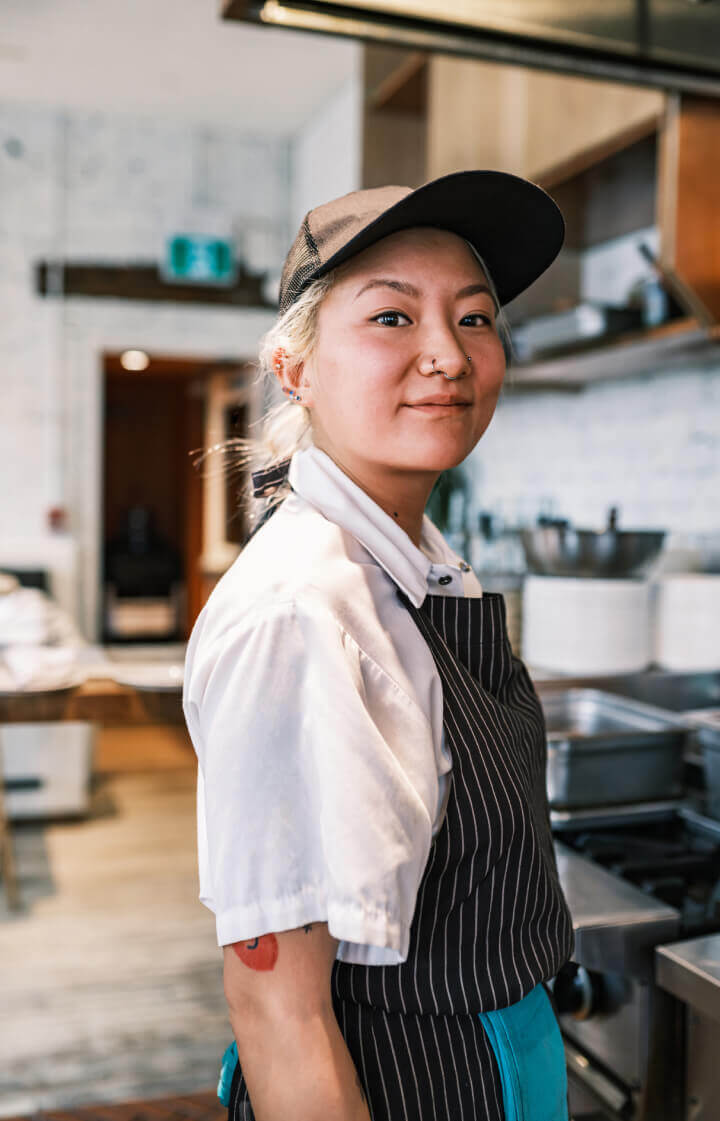 A young Asian woman with a nose ring and baseball hat stands in a restaurant kitchen