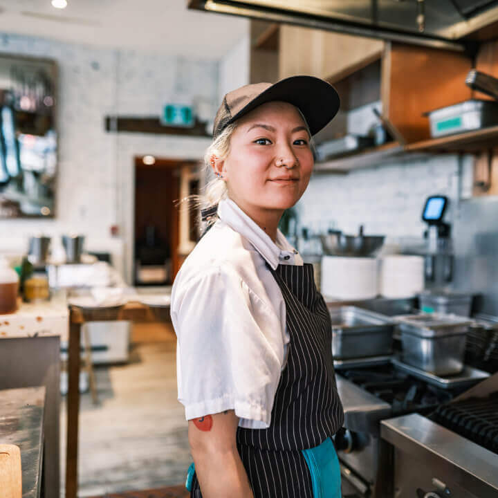 a young woman with a nose ring and baseball hat stands in a restaurant kitchent