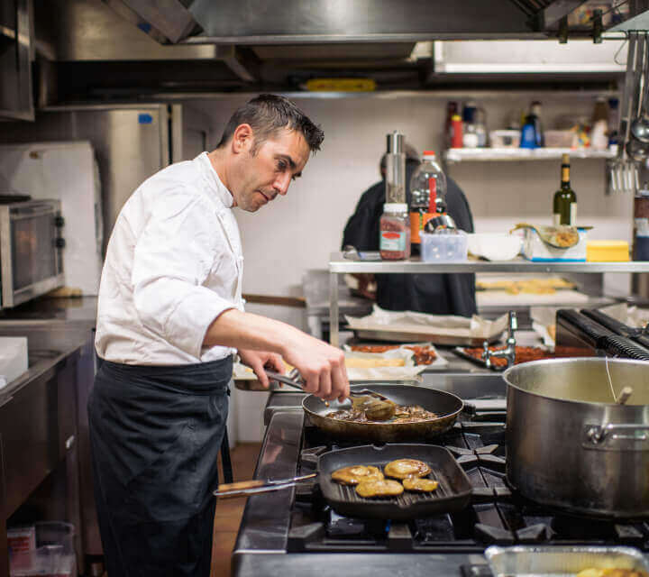 A male chef is pan sautéing steak in a commercial restaurant kitchen