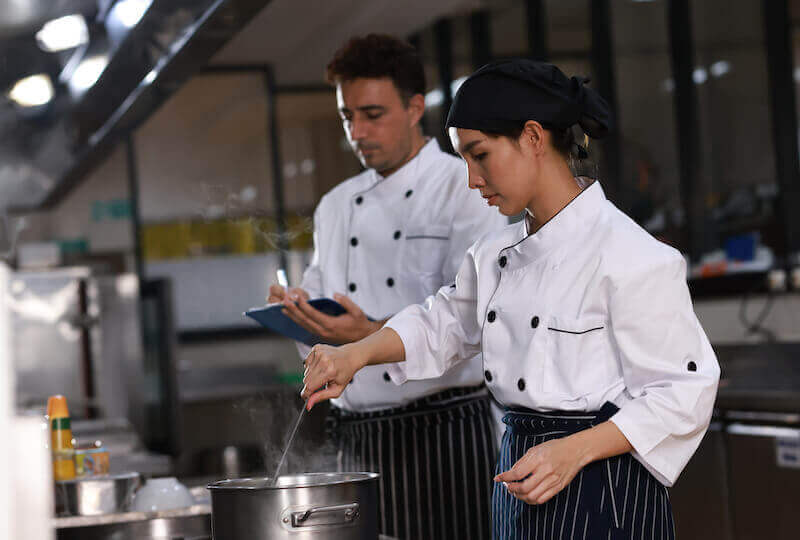  A chef is taking notes while a female cook is stirring a pot of soup in a restaurant kitchen.