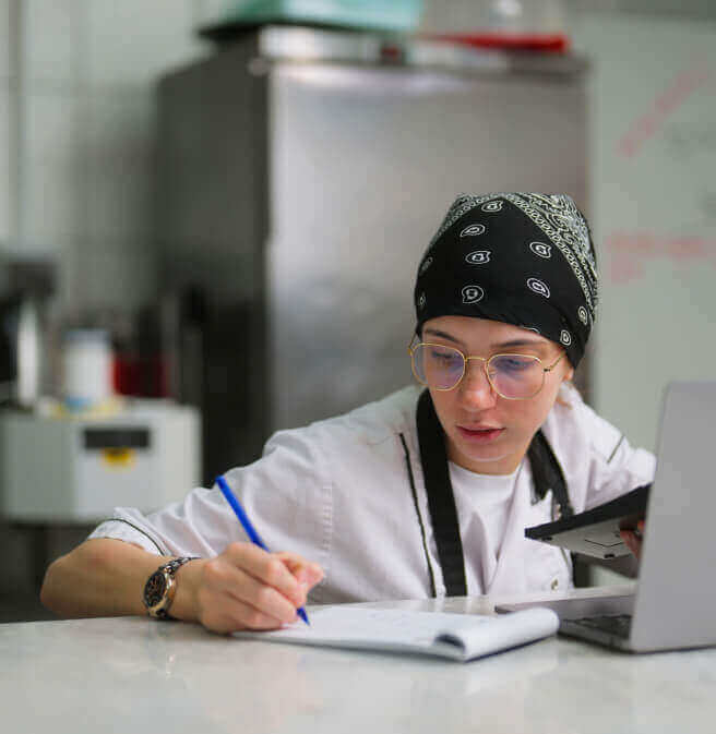 Young female chef writing notes on a notepad in front of her laptop computer in a commercial kitchen