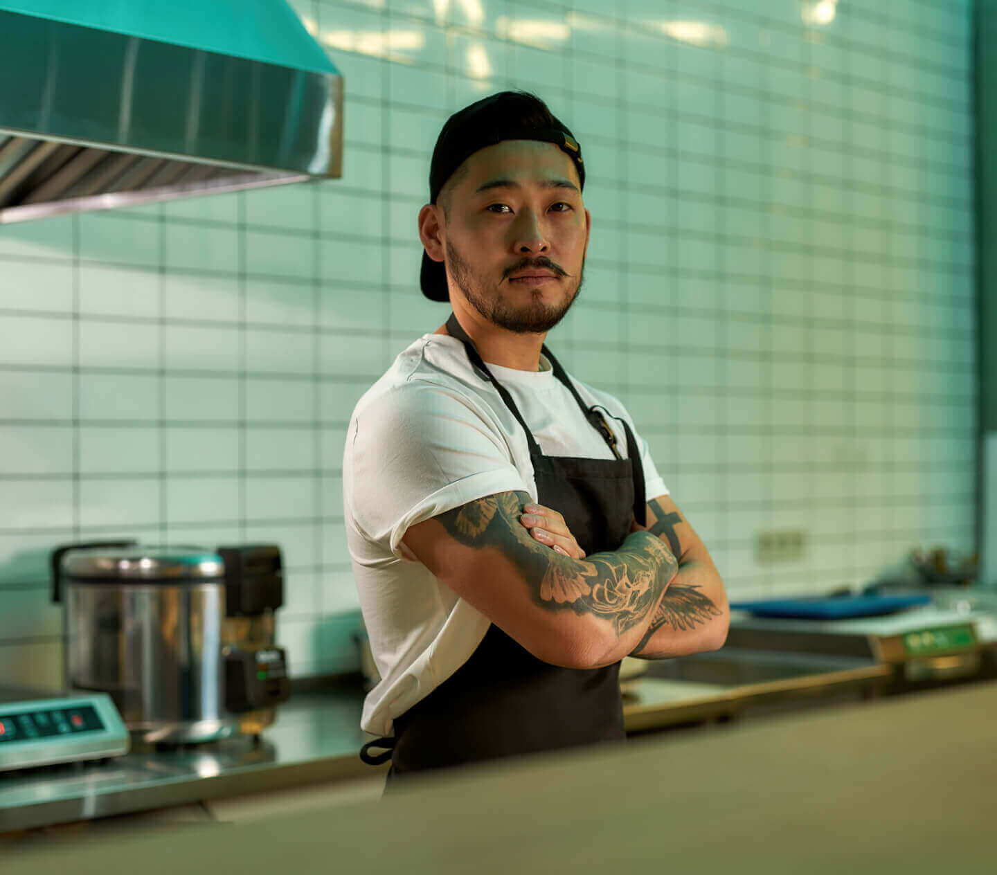 An Asian chef standing with his tattooed arms folded in a restaurant kitchen