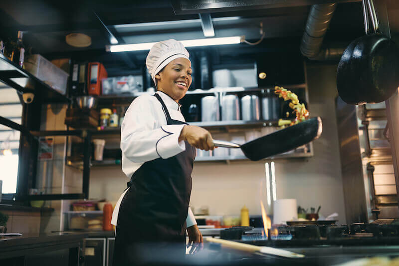 A chef in a black apron tosses a stir-fry in a professional kitchen.