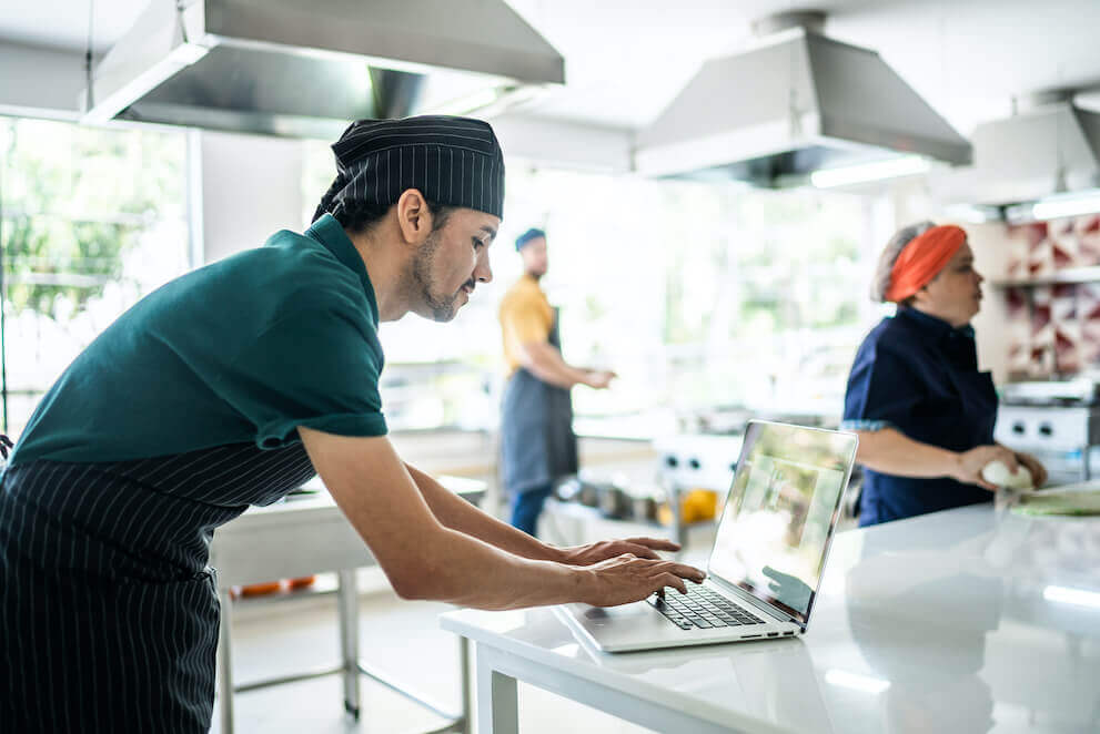 A chef uses his laptop in the midst of a brightly lit kitchen with two other chefs in the background.