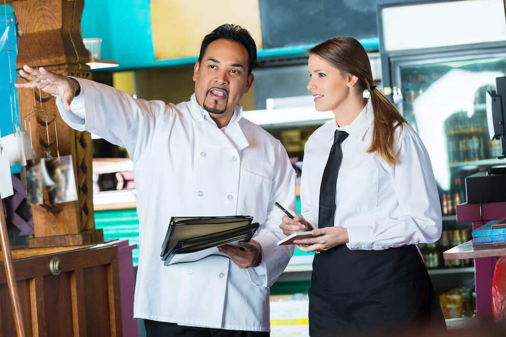 Person in a chef's uniform holding a stack of menus gestures toward something off screen, while a restaurant employee holds a notepad and pen.