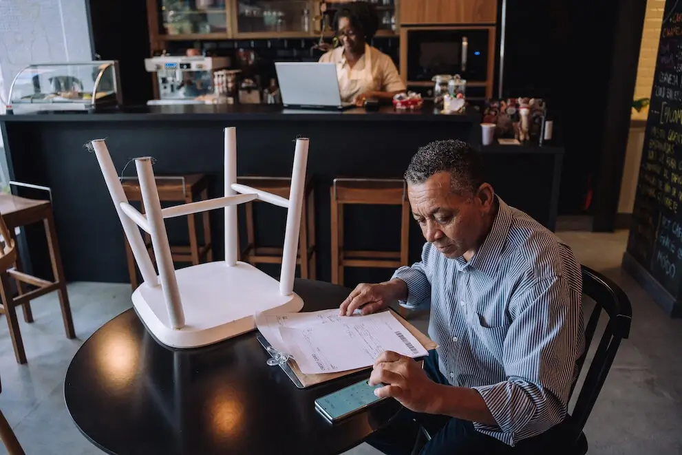 A restaurant owner sits at a cafe table after hours with a clipboard and a calculator, while another person in the background sits at a counter with their laptop open