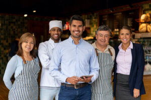 A restaurant owner stands holding a tablet with a group of people around them, all facing the camera and smiling.