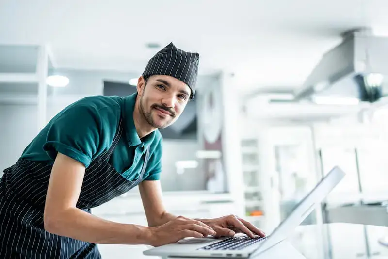 Person in chef's hat and apron smiles while working on laptop in a professional kitchen.