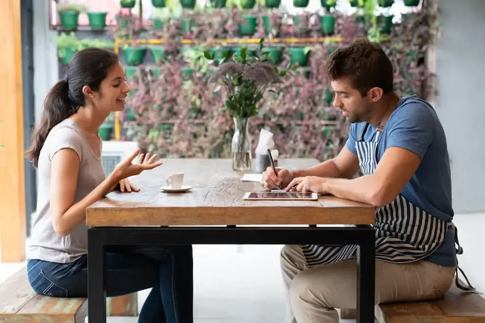A person in an apron takes notes while interviewing a job candidate in a restaurant setting.