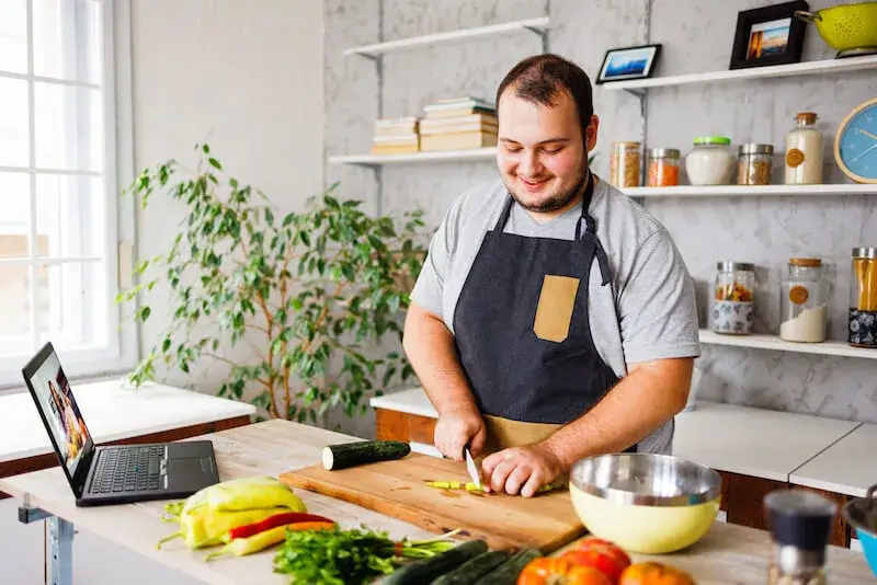 A person smiling in an apron, chopping vegetables in a personal kitchen while an instructional video plays on their laptop.
