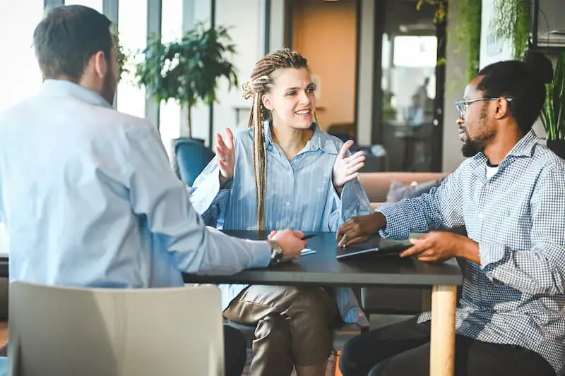 Three people sit at a table in button-down shirts having a discussion while two of the people take notes. 