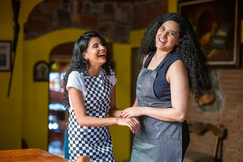 Two young people in aprons, laughing and smiling in a dining room setting. 