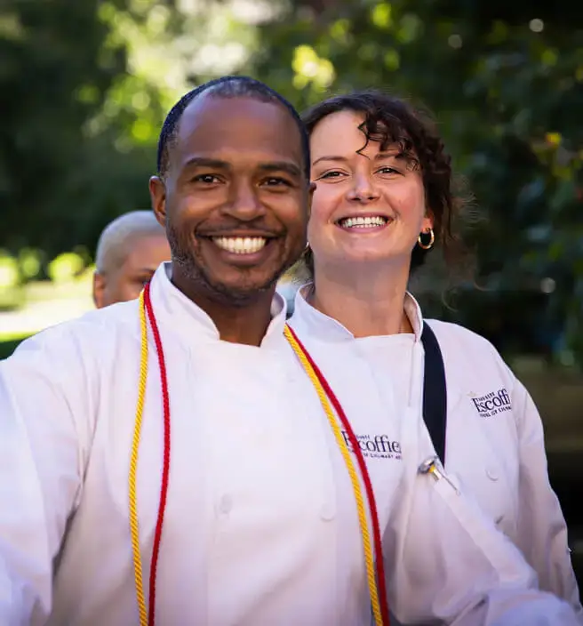 Two smiling Escoffier culinary graduates in Escoffier chef coats pose outside during graduation day