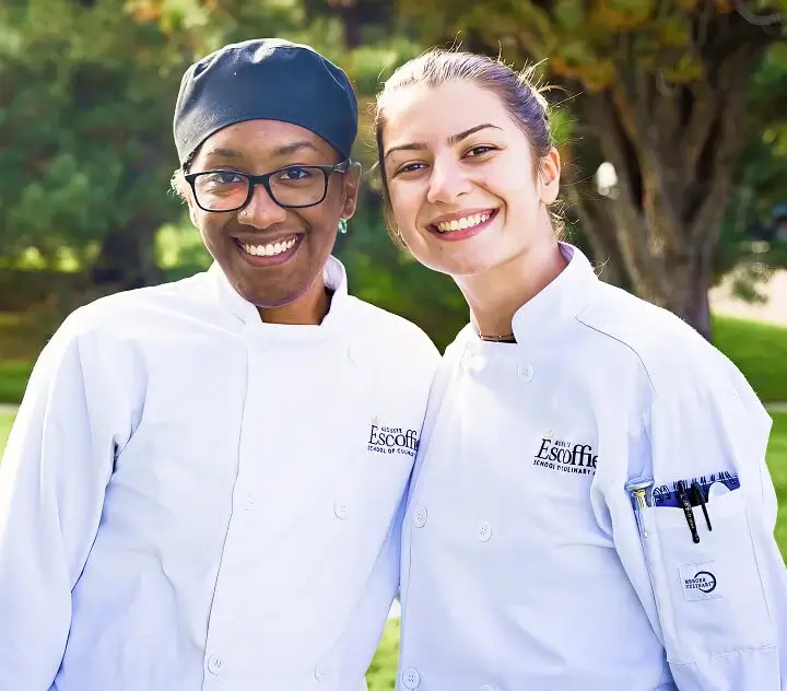 Two female Escoffier culinary students in Escoffier chef uniforms smile together outside on campus