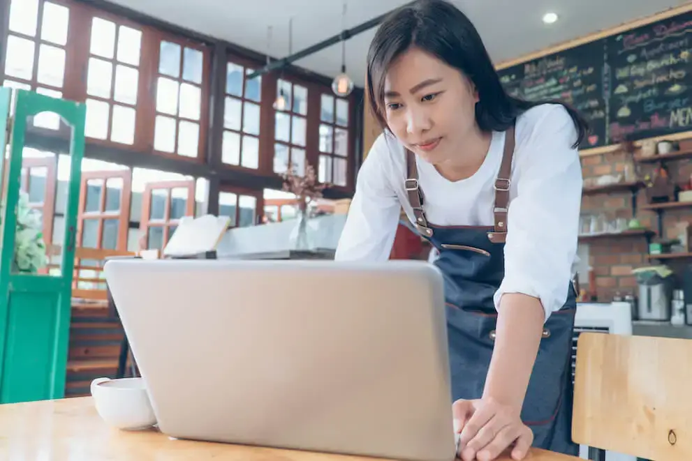 A person wearing an apron stands at a table in a brightly lit cafe looking at a laptop.