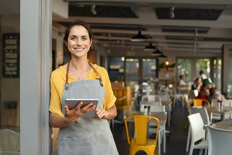 A person in an apron smiles and looks at the camera while holding a tablet and standing in the open door to a restaurant dining room.