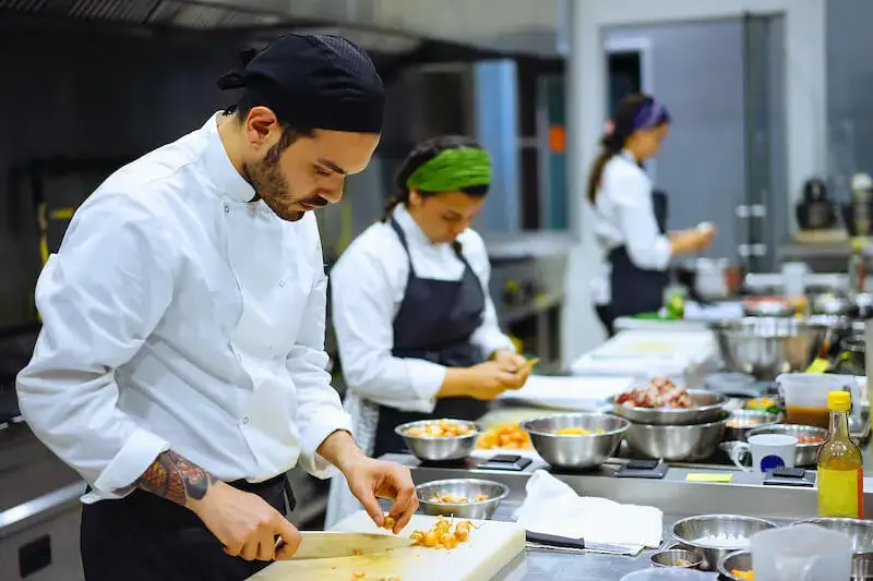 Several people in chef’s uniforms work side by side at a counter covered with bowls, cutting boards, vegetables, and other culinary items in a restaurant kitchen.