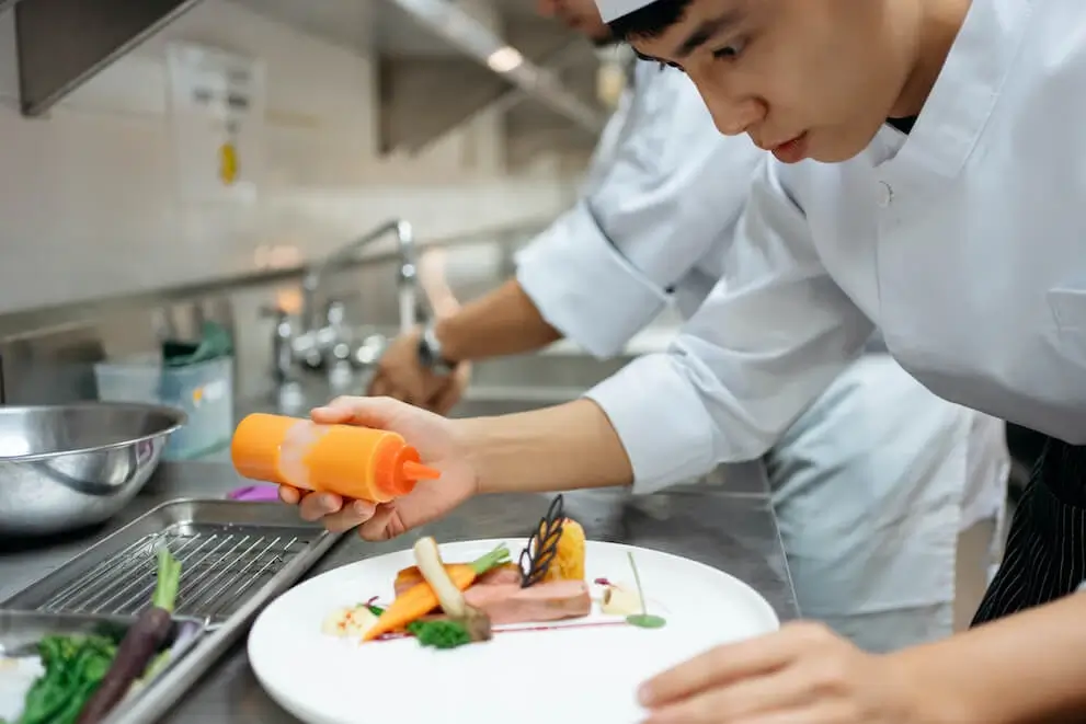Young chef plating dish in professional restaurant kitchen.