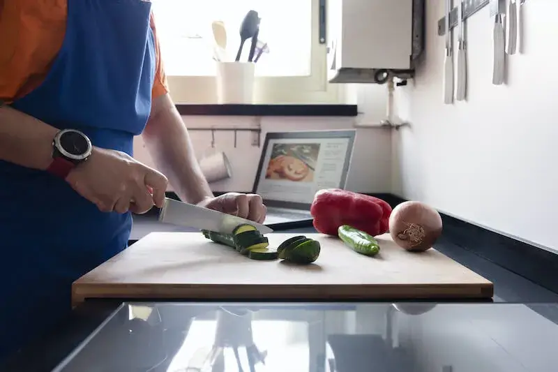 Man chopping vegetables in a home kitchen, with a laptop in the background whose screen shows chopped vegetables.