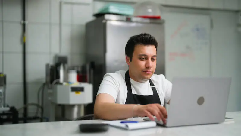 A person in a chef’s apron sits at a metal counter in a restaurant kitchen using a laptop with an open notebook and calculator nearby.
