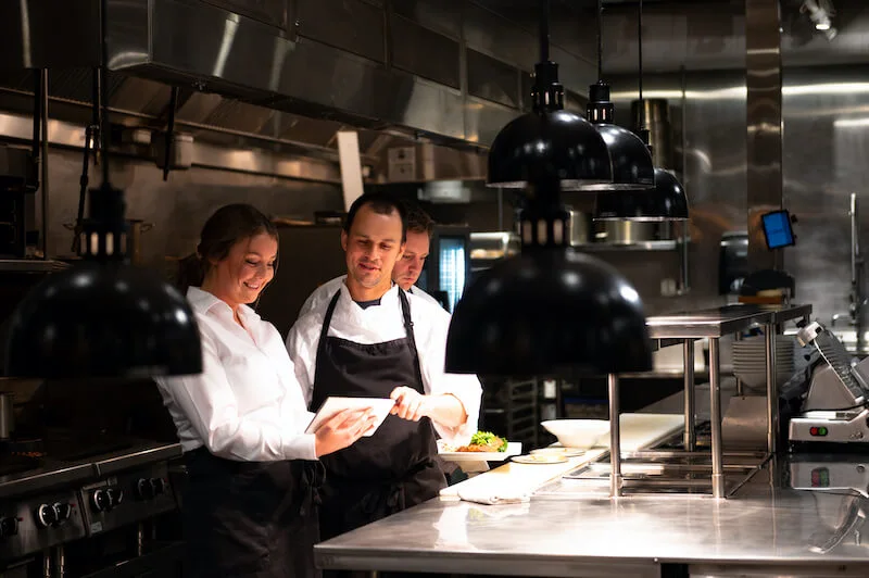 A person wearing a chef’s uniform and a person wearing a server’s uniform stand in a restaurant kitchen smiling and looking at a tablet while another person holds plated dishes in the background.
