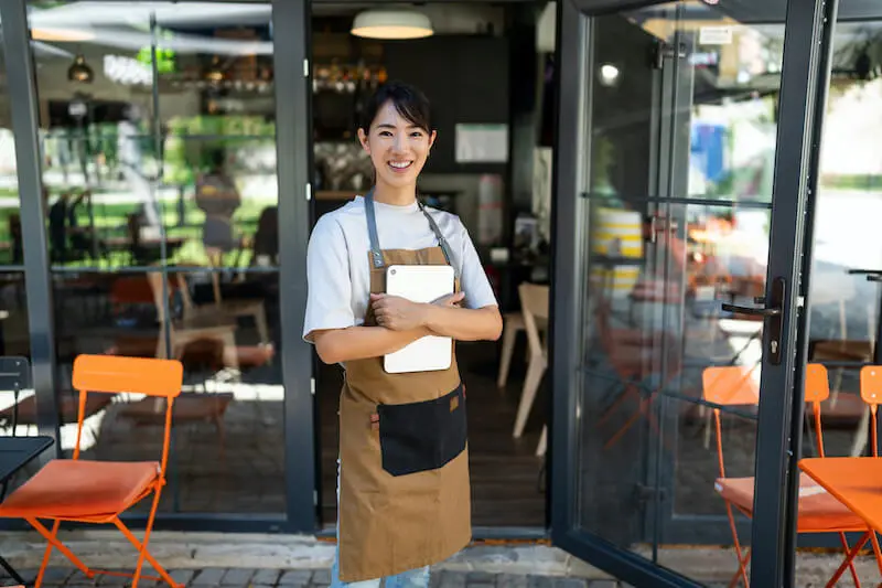A smiling person wearing a server’s uniform stands with arms crossed holding a tablet on a patio outside the open door to a café.