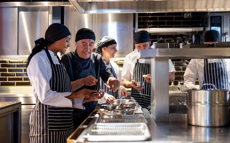 An older person in an executive chef’s uniform gives advice to a younger person in a chef’s uniform holding a mushroom at a workstation covered with several metal bowls in a restaurant kitchen while several other people in chef’s uniforms work behind them.