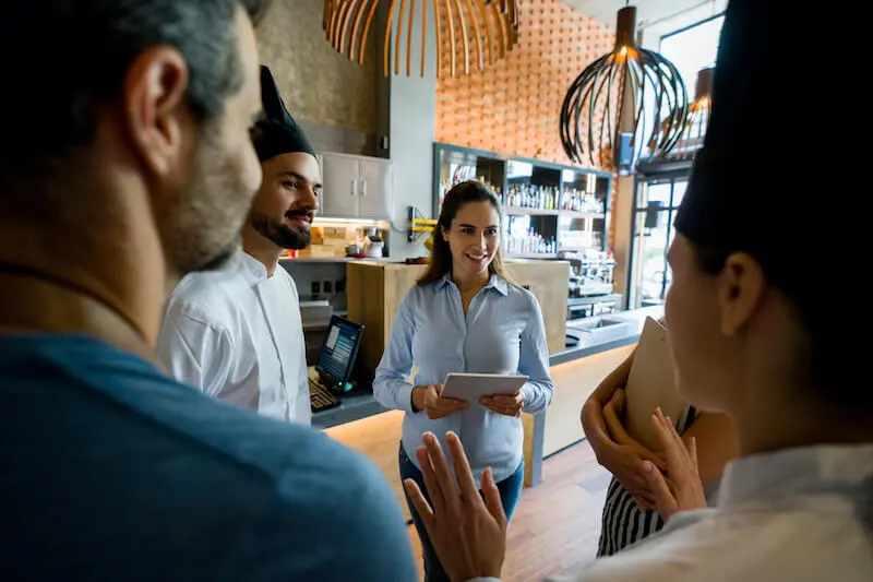 A person in casual business attire smiles and holds a tablet as they listen to a person wearing a chef’s apron while three other people listen in a modern restaurant dining room.