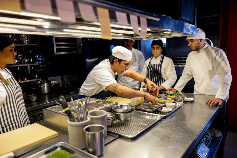 A person wearing a chef’s uniform reaches over a metal work table toward several dishes of ingredients in a restaurant kitchen while four other people in chef’s uniforms watch.
