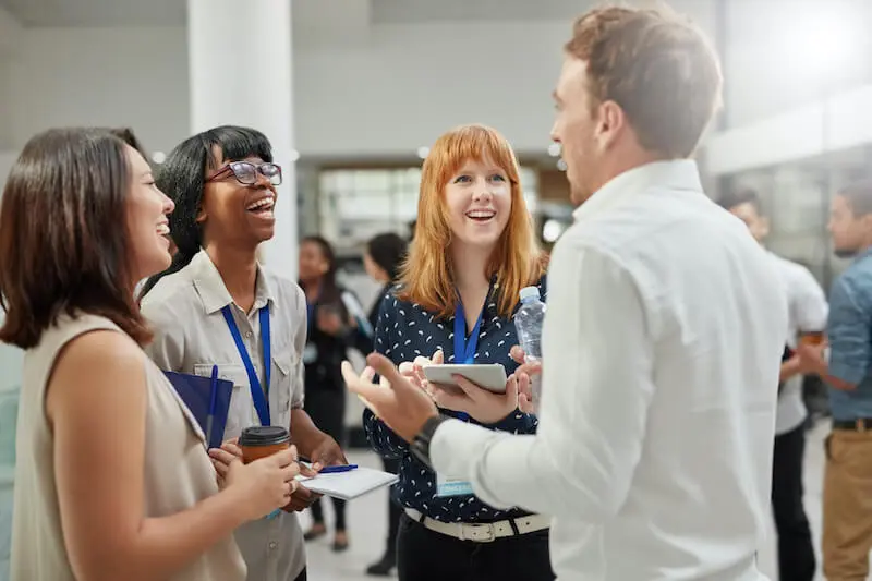 Several people in casual business dress and wearing conference lanyards stand in a circle talking and laughing in a crowded event space.