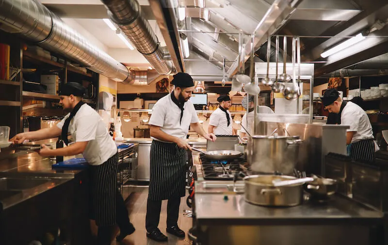 Several people in chef’s uniforms and head coverings work at different stations in a clean, orderly restaurant kitchen.