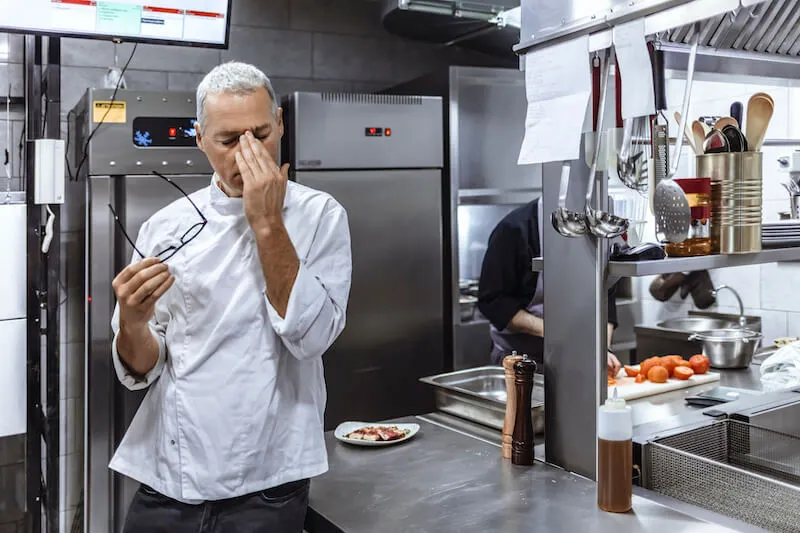 A chef in a commercial kitchen stands with eyes closed and hand on face, appearing exhausted and overwhelmed.
