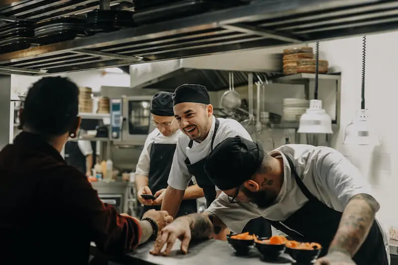 A group of kitchen staff laugh and work together in a high-end restaurant kitchen during service.