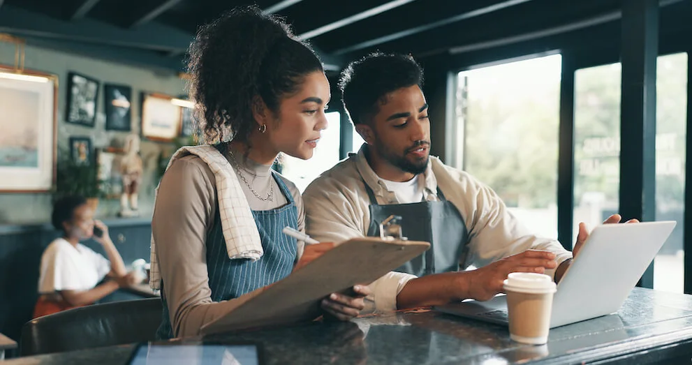 A person wearing an apron with a dish towel over their shoulder stands at the counter of a coffee shop writing on a clipboard next to another person wearing an apron seated at the counter using a laptop.