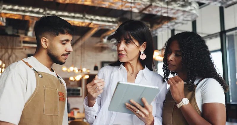 A restaurant manager holding a tablet briefs two kitchen staff members in an industrial restaurant setting. The manager is clearly communicating information from the digital device.