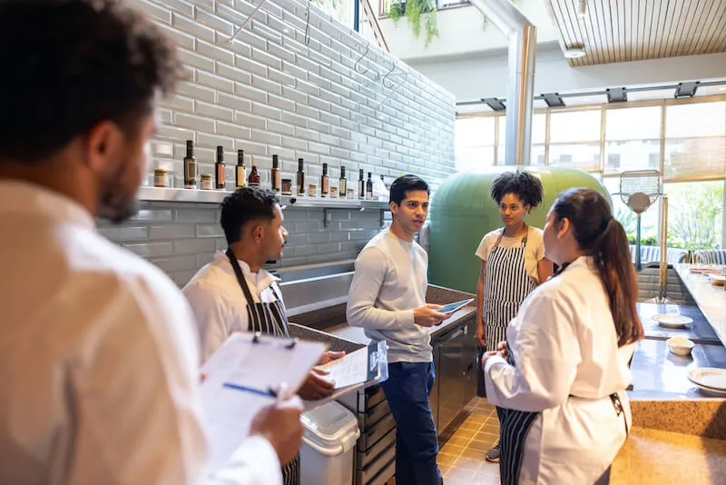 Restaurant owner leading a staff meeting with a small group of employees in a restaurant dining area.